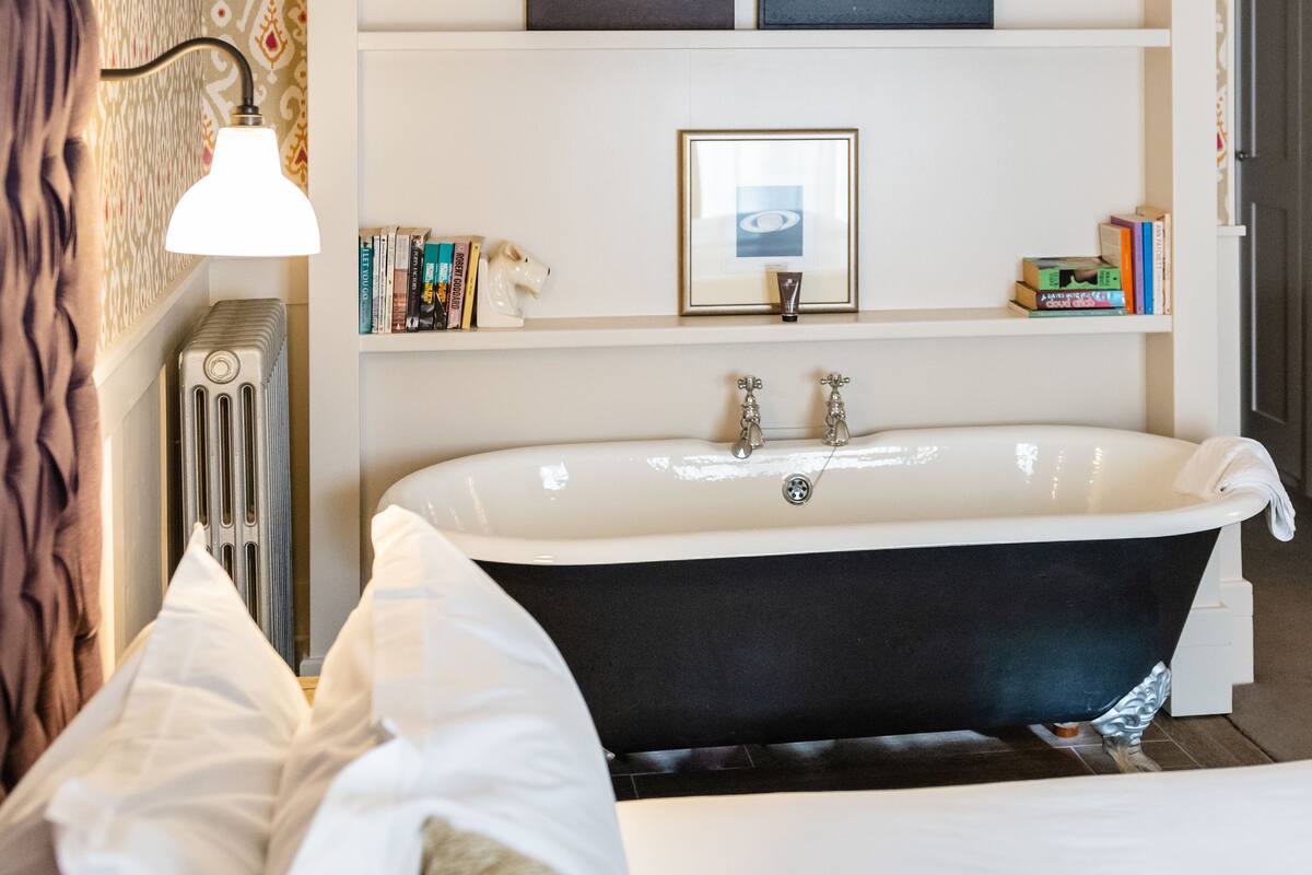 A bathroom with a freestanding bathtub, a radiator, a shelf with books, and framed artwork.