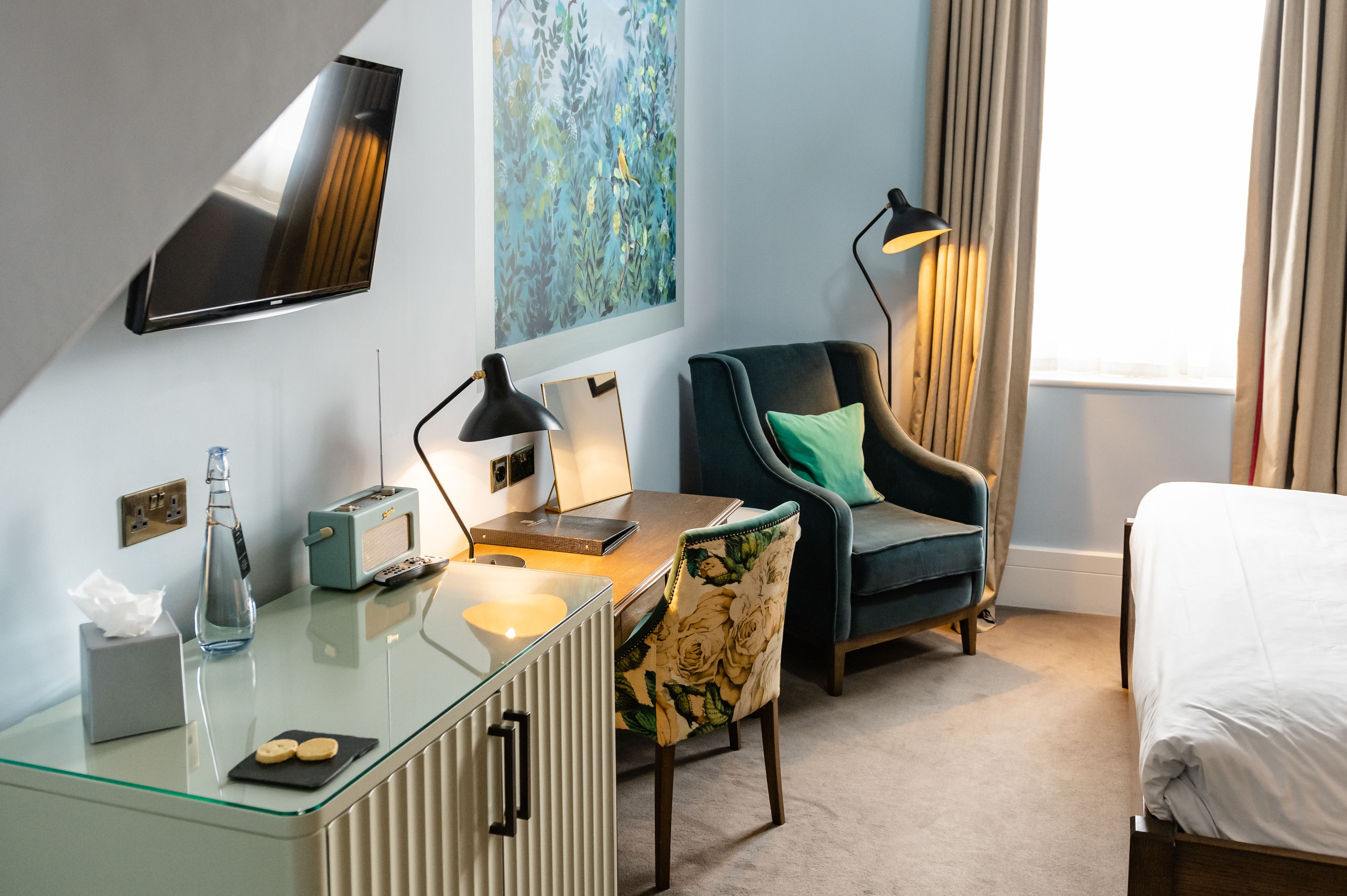 Une chambre d’hôtel avec un bureau en verre, une chaise, un lit, un téléviseur fixé au mur et une fenêtre avec des rideaux.