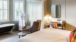 Habitación de hotel con una cama grande, un área de descanso con una silla, un tocador con espejo, una bañera y una ventana con persianas.
