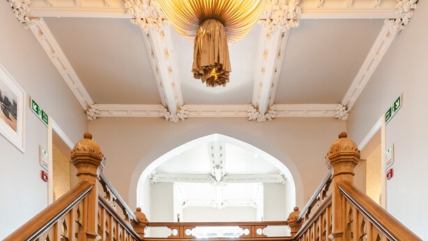 Una imponente escalera con detalles de madera ornamentales y un candelabro, que conduce a un pasillo luminoso con piso de madera.