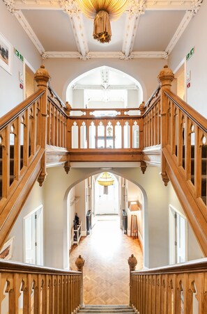 A grand staircase with ornate woodwork and a chandelier, leading to a bright hallway with a wooden floor. - Didsbury House Hotel (Manchester)