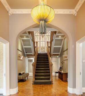 A grand staircase with a chandelier, a wooden bench, and a candelabra in a spacious hallway with a wooden floor. - Didsbury House Hotel (Manchester)