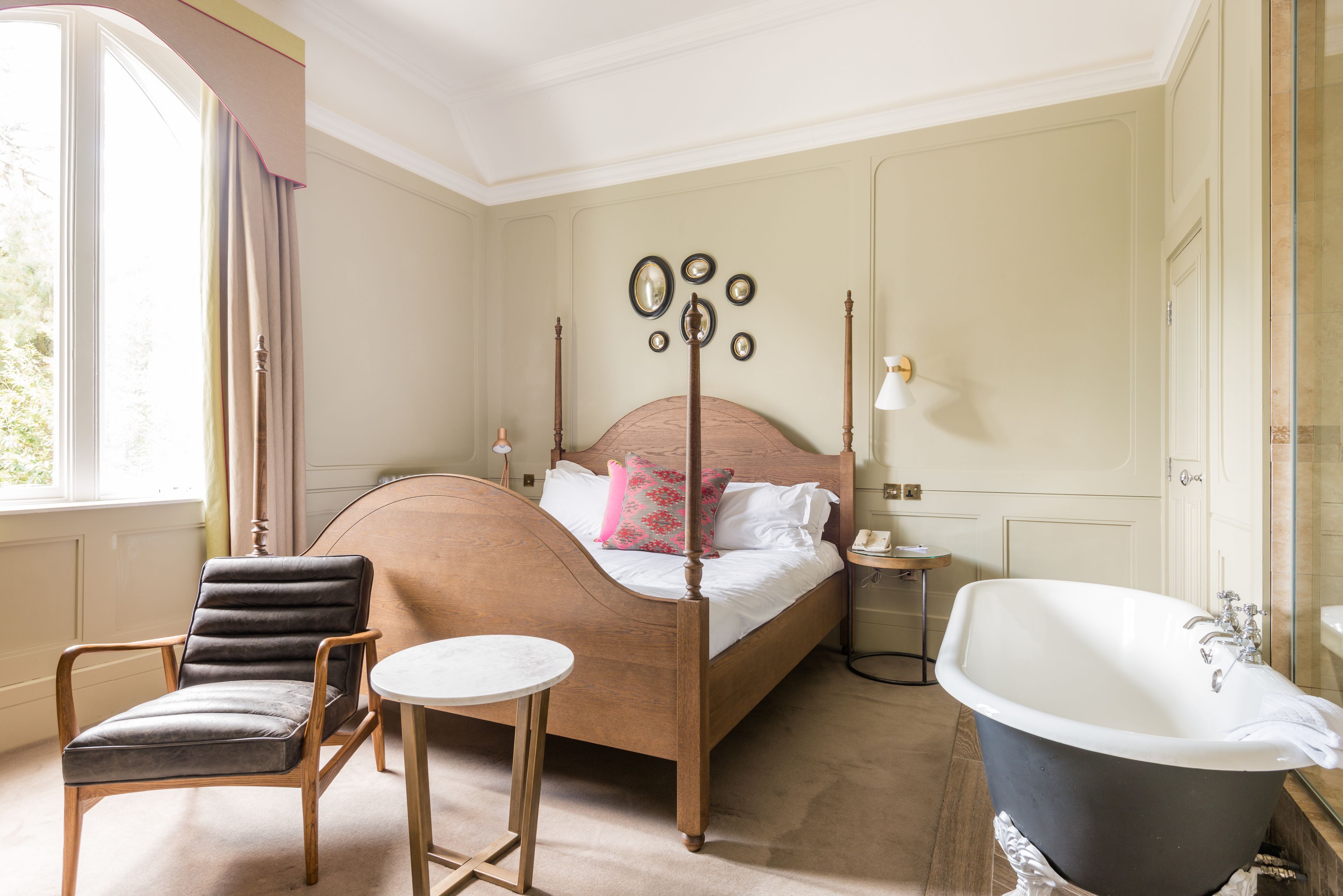 Un dormitorio con cama con dosel, un sillón, una mesita redonda, una bañera grande y una mesita de noche con lámpara.