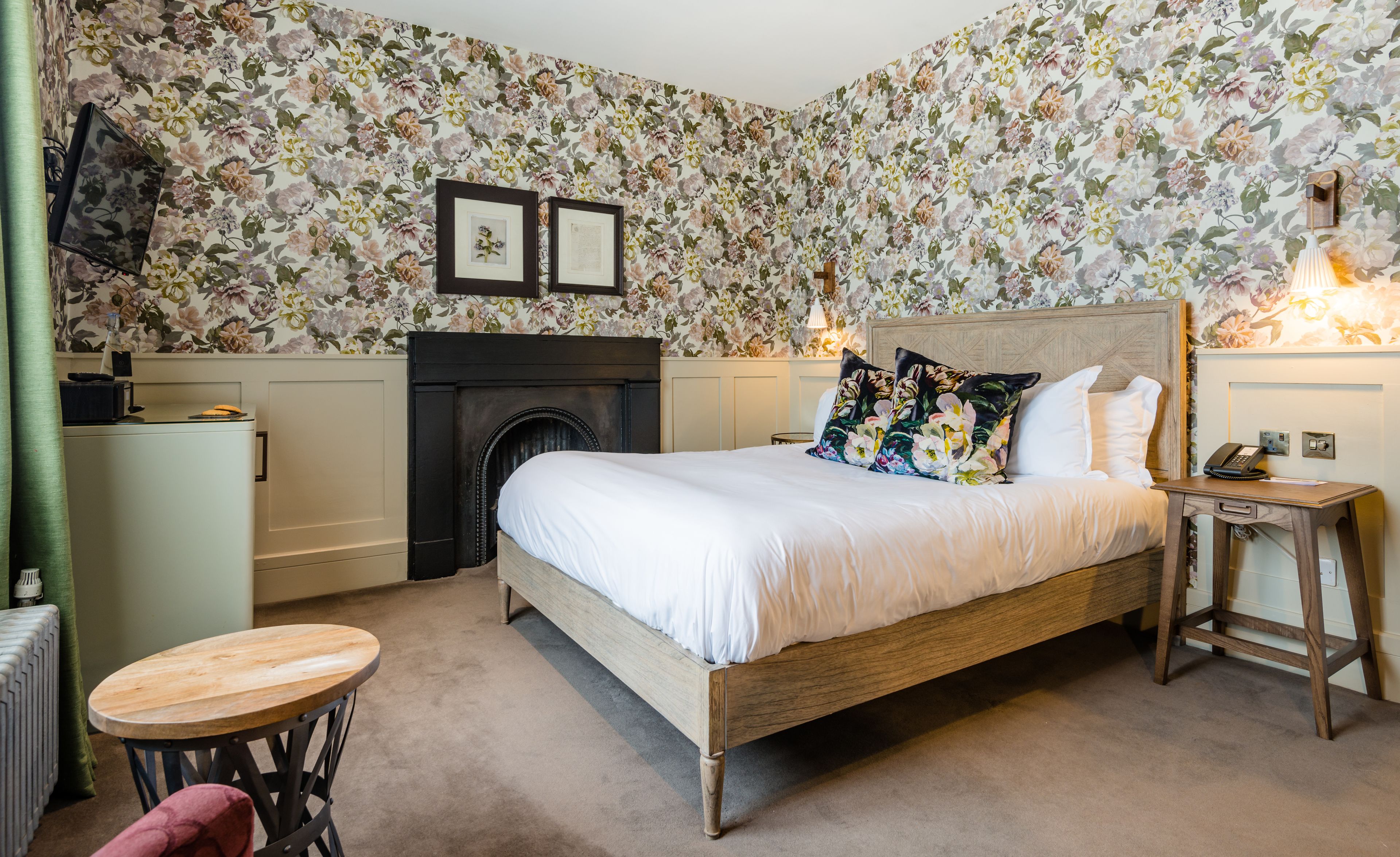 Een slaapkamer met een bed, een tafeltje, een stoel, een open haard en een wand met bloemenbehang.