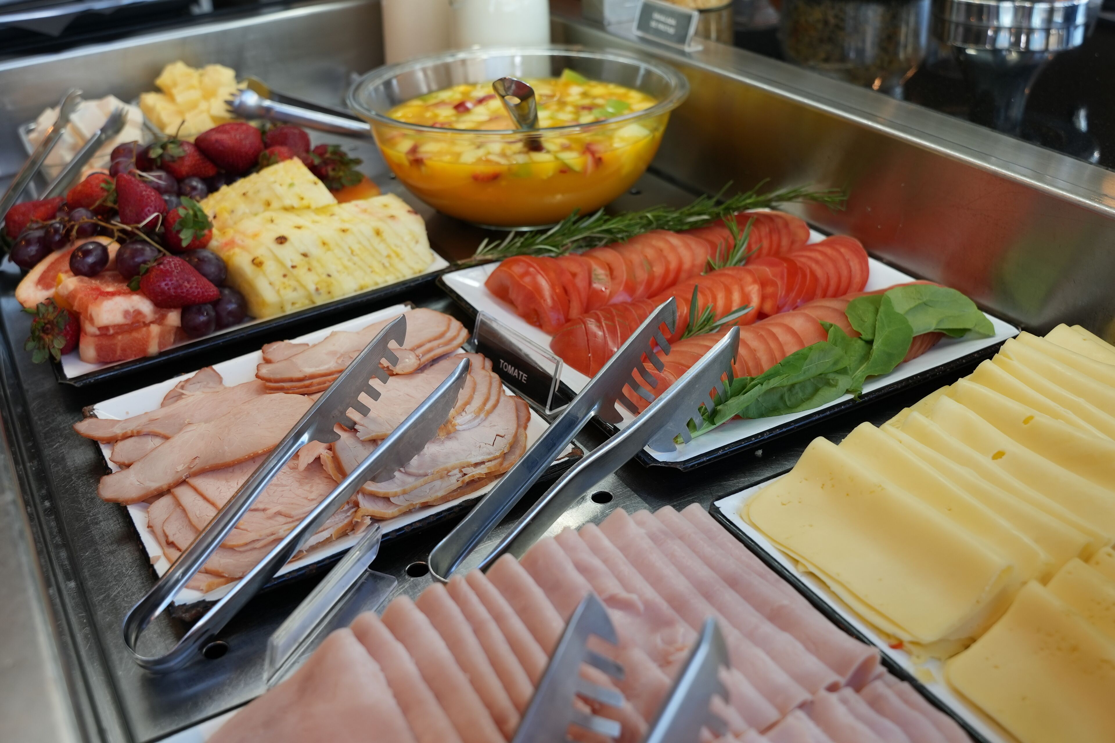 A buffet with various food items including sliced meats, cheeses, fruits, and a bowl of fruit salad.