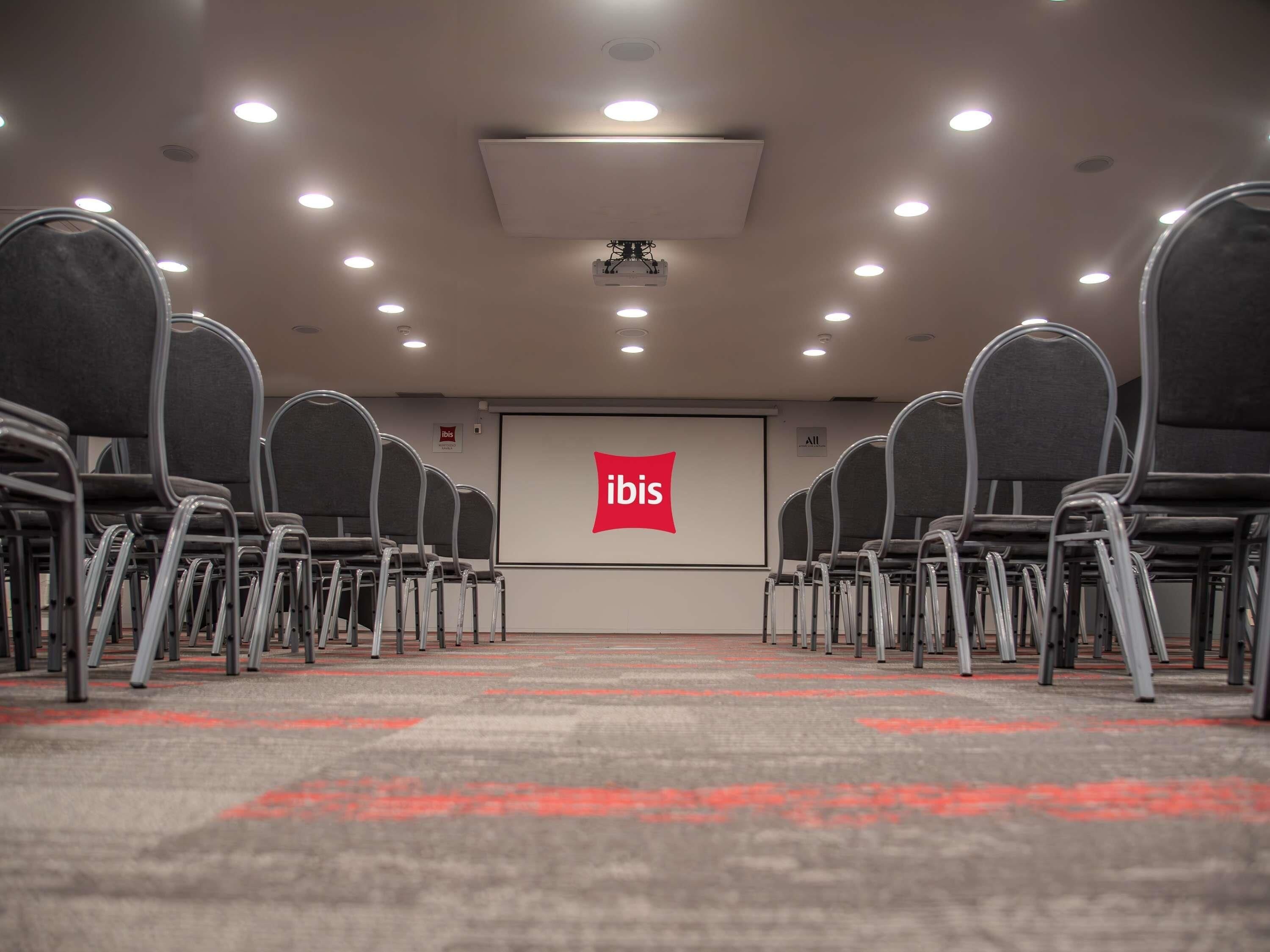 A conference room with rows of chairs, a projector screen with the ibis logo, and ceiling lights.