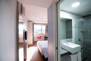 A modern hotel room with a bed, a desk, a red chair, a large window, and a bathroom with a sink and shower. - Hotel ibis Montevideo Rambla (Montevideo)
