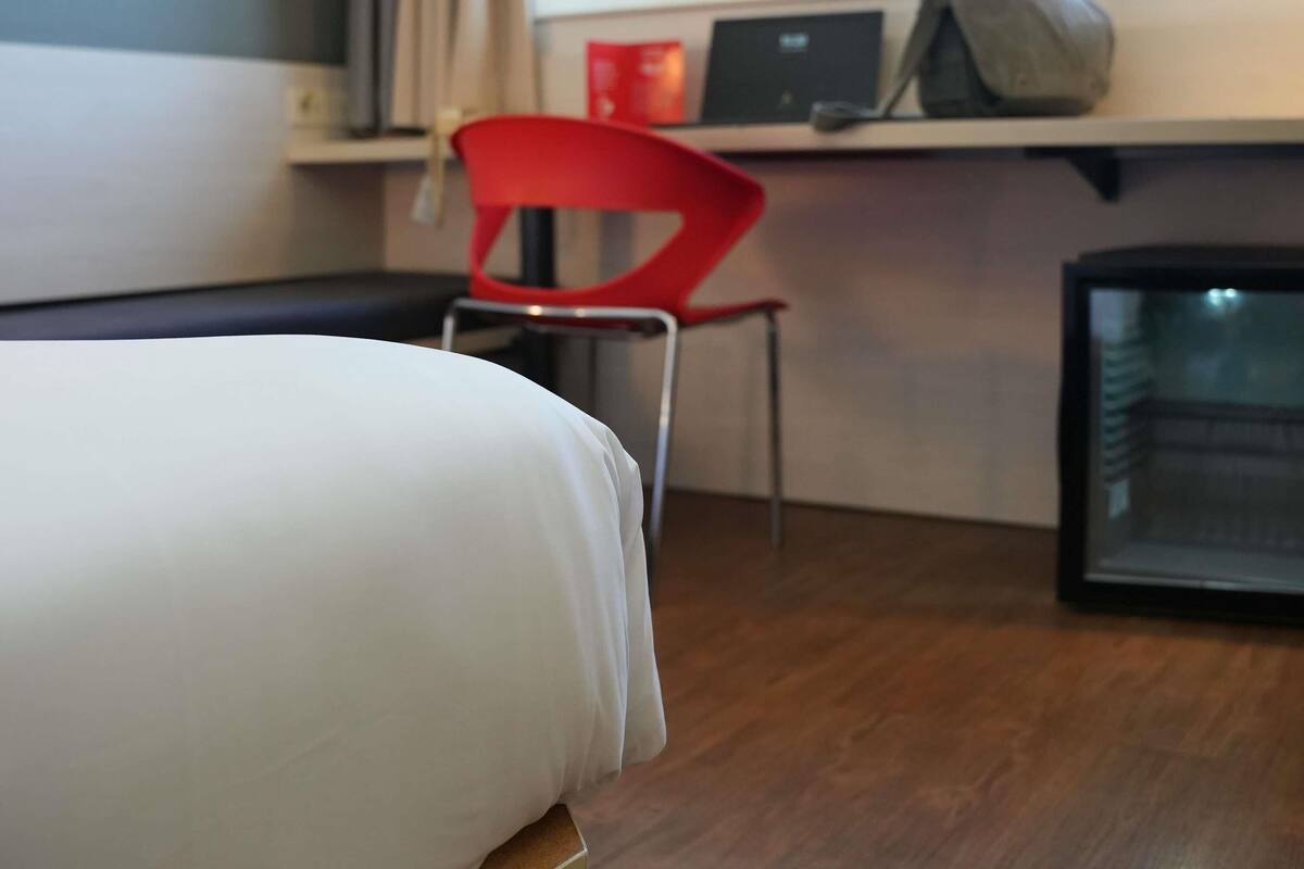 A hotel room with a bed, a red chair, a desk with a computer, and a small refrigerator.