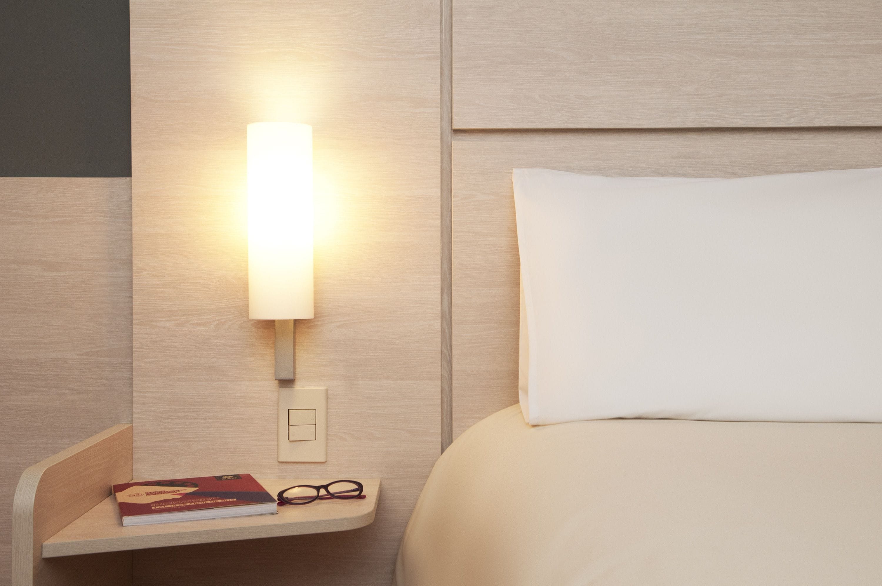 Una cama bien hecha con un almohadón blanco y una mesita de noche con un libro y unos lentes.