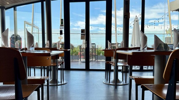 Un interior moderno con ventanas amplias, mesas y sillas de madera, que ofrece vistas al mar.