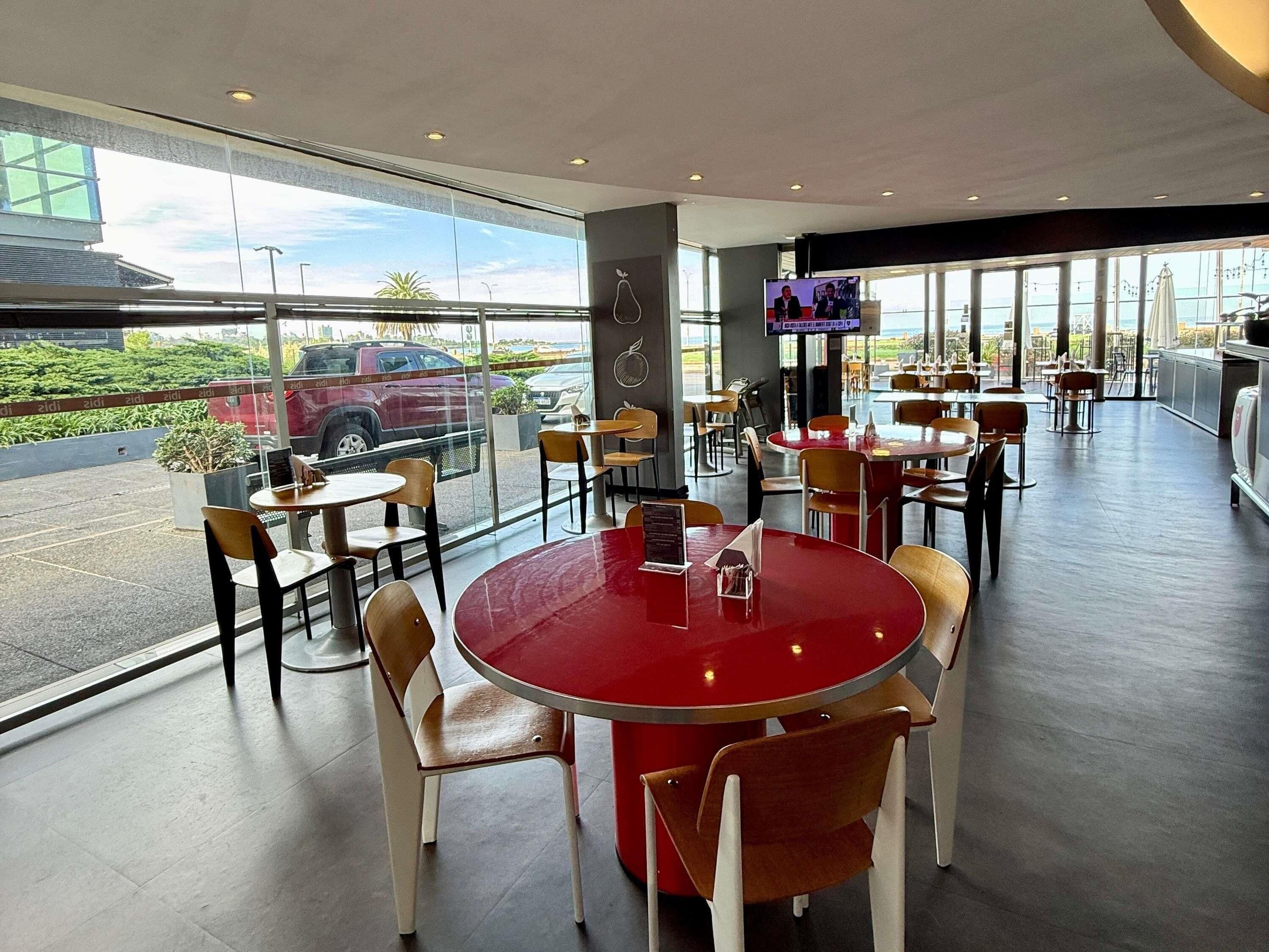 Een modern restaurant met ronde tafels en stoelen, grote ramen en uitzicht op de parkeerplaats.