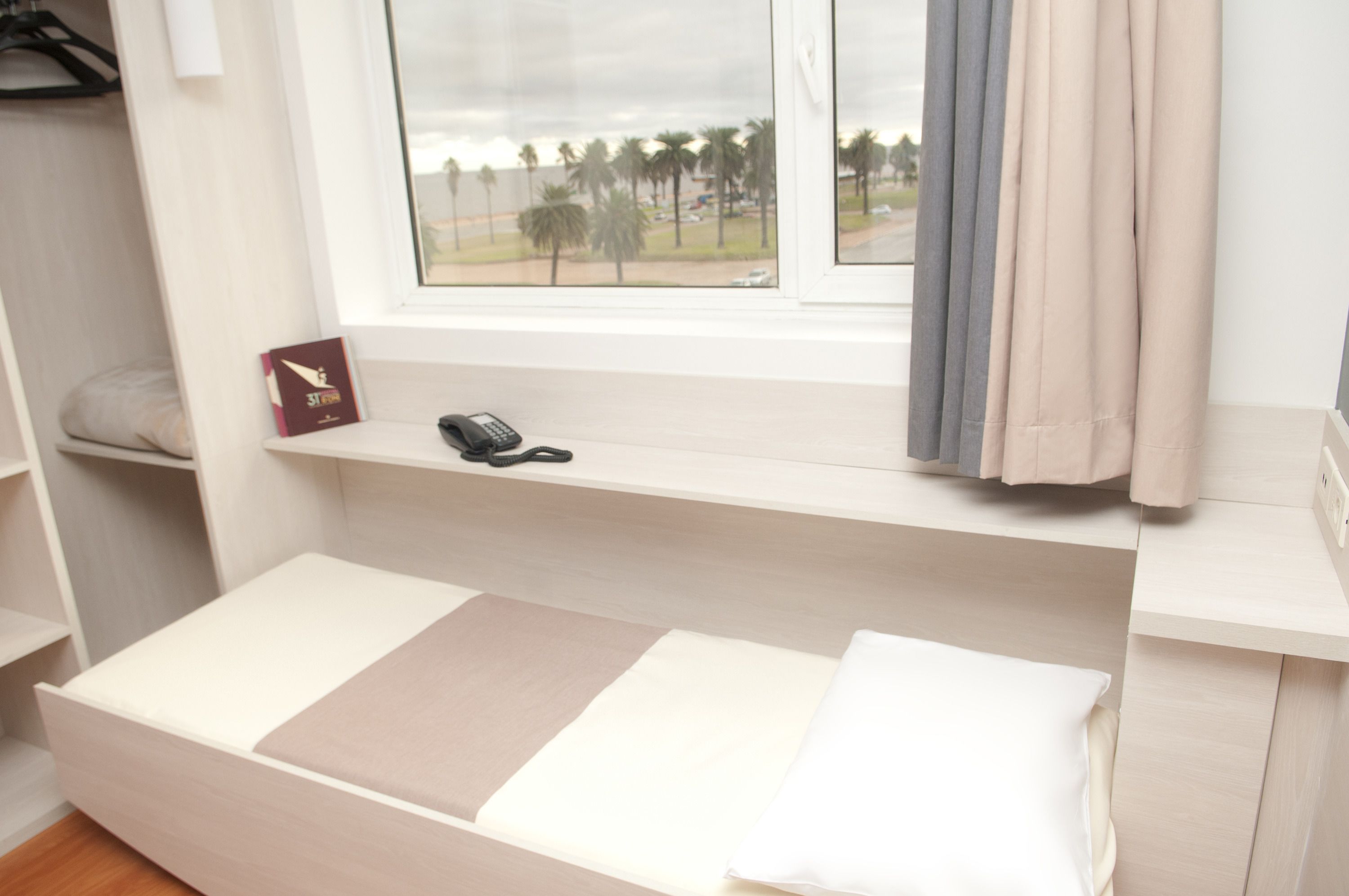 Een slaapkamer met een enkel bed, een raam met gordijnen, een nachtkastje en een klein plankje met een boek en een telefoon.