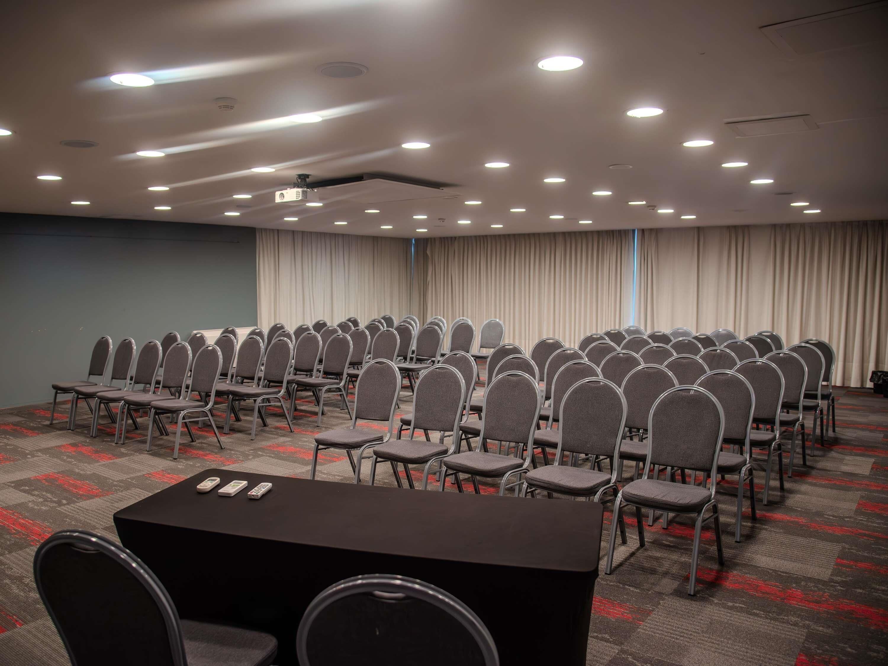 A conference room with rows of chairs, a black table with remotes, and a projector on the ceiling.
