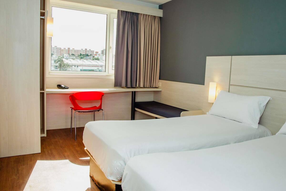 A hotel room with two beds, a desk with a chair, a window with curtains, and a view of buildings outside.