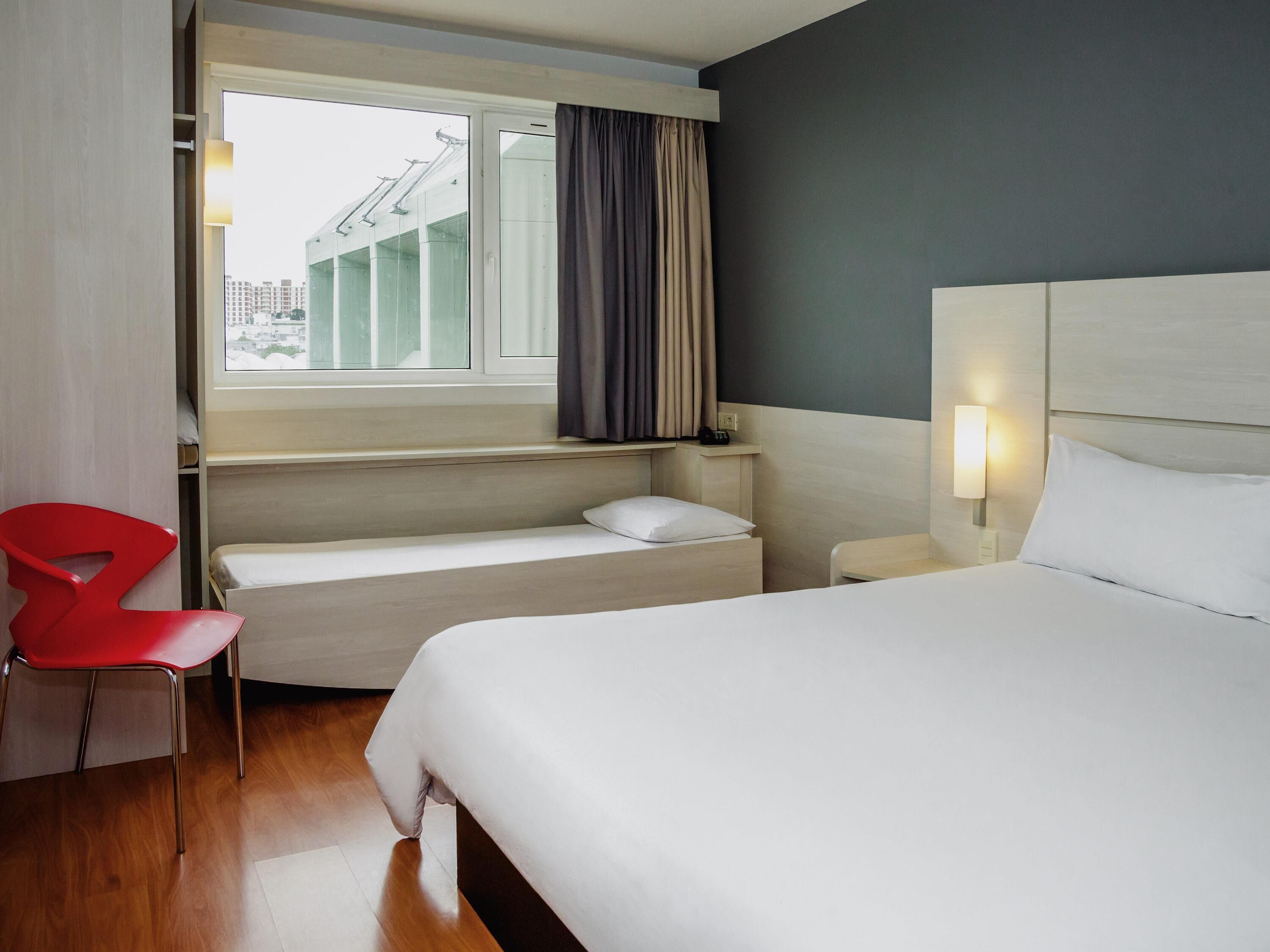 A hotel room with a bed, a red chair, a small table, a nightstand, and a window with curtains.
