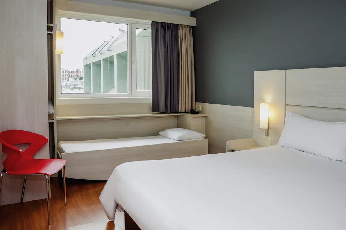 A hotel room with a bed, a red chair, a small table, a nightstand, and a window with curtains.