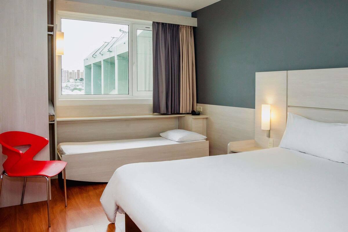 A hotel room with a bed, a small desk, a chair, a window with curtains, and a view of buildings outside.