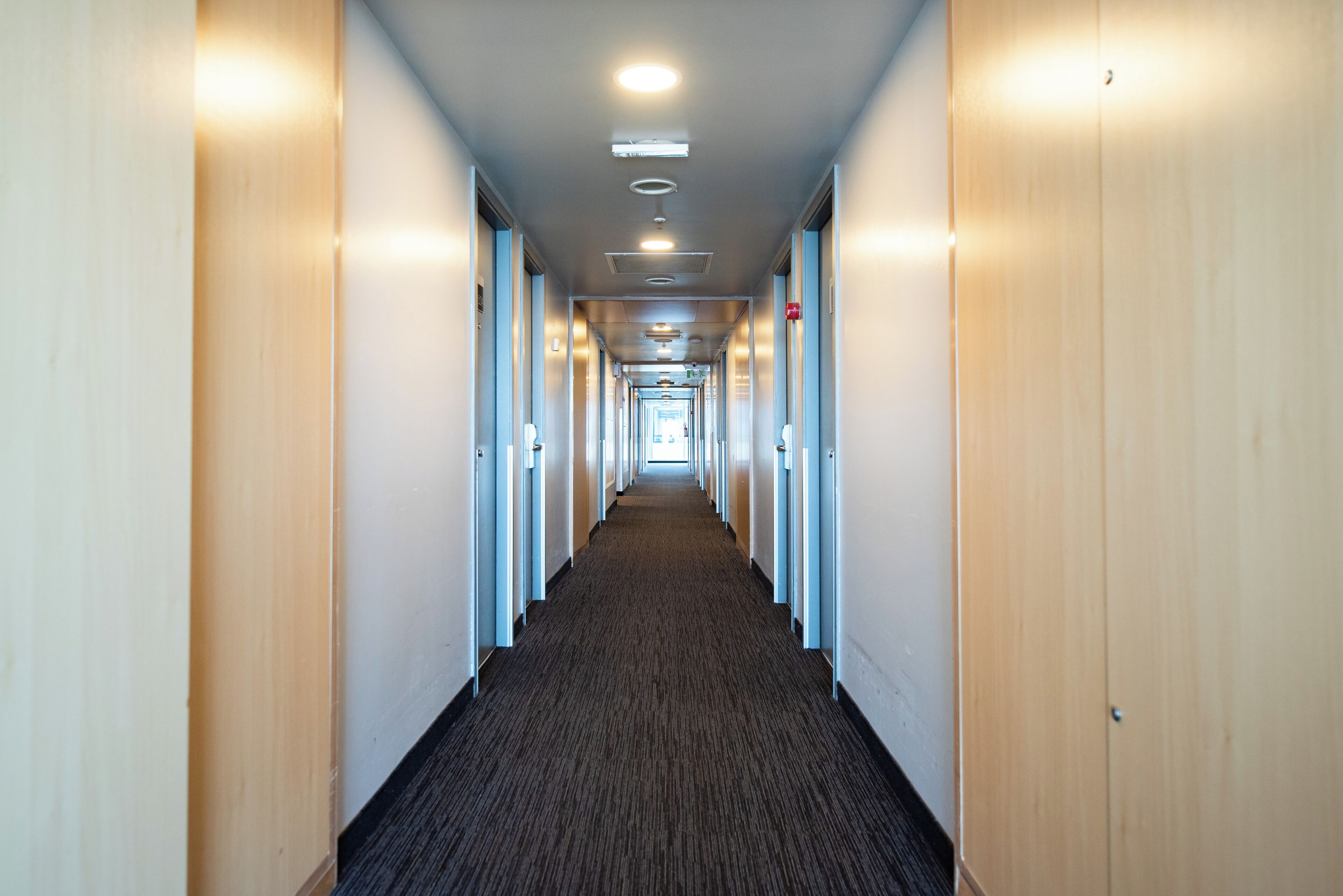 A long, narrow hallway with doors on both sides, leading to a bright room at the end.