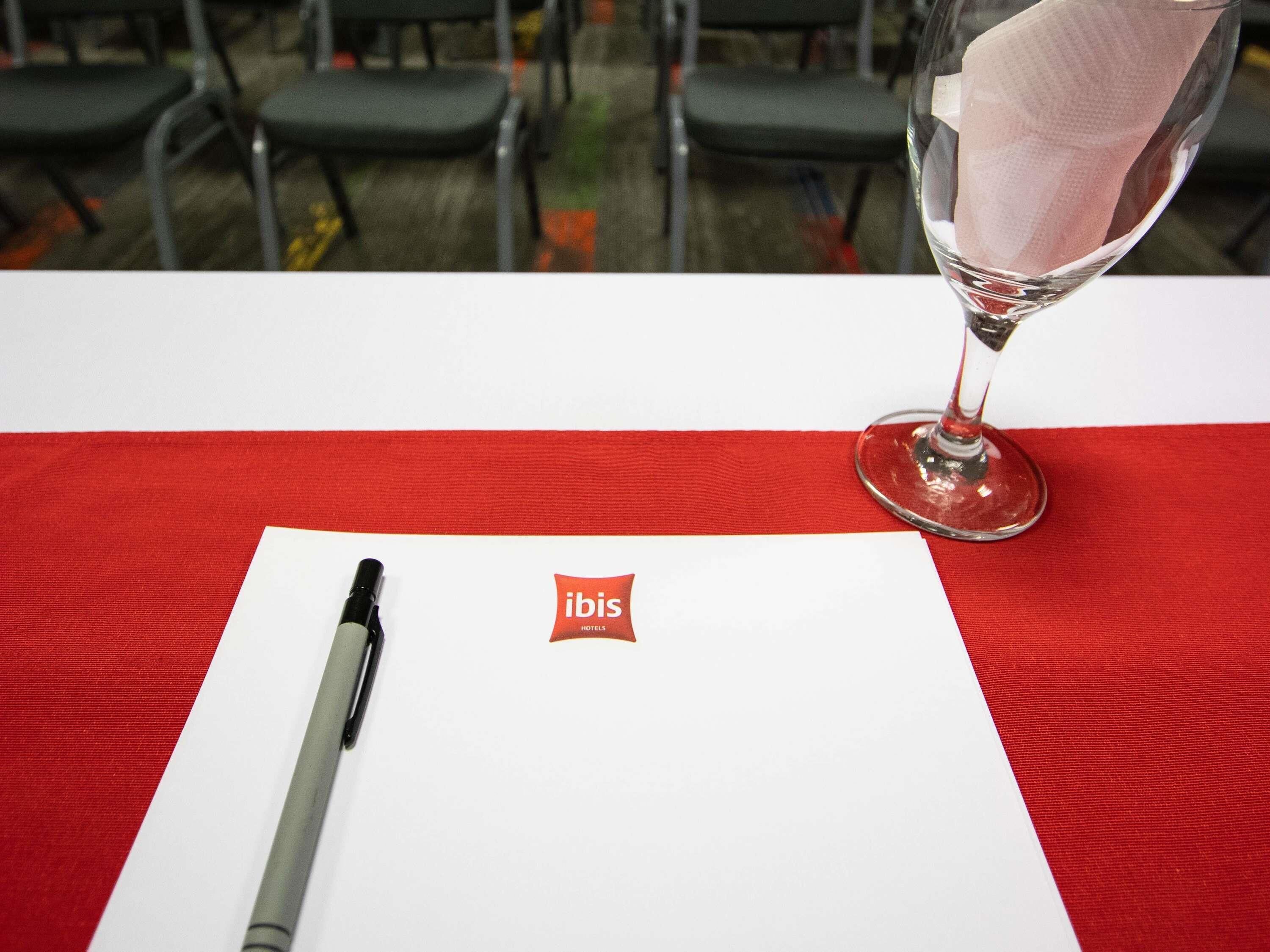 Una mesa con mantel rojo, un bolígrafo, un vaso con servilleta y un papel con el logotipo de 'ibis Hotels'.