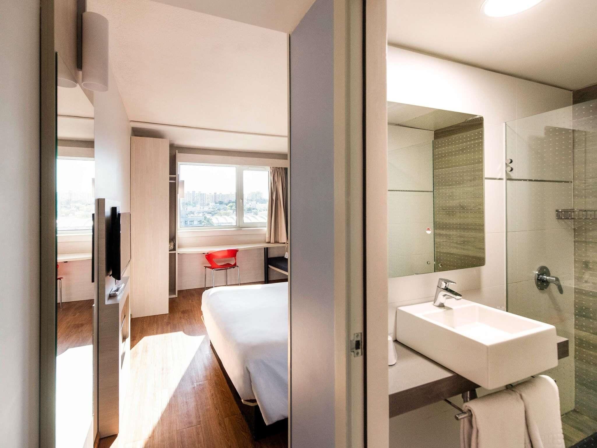 A modern hotel room with a bed, a desk with a chair, a large window, and a glass-enclosed shower area.
