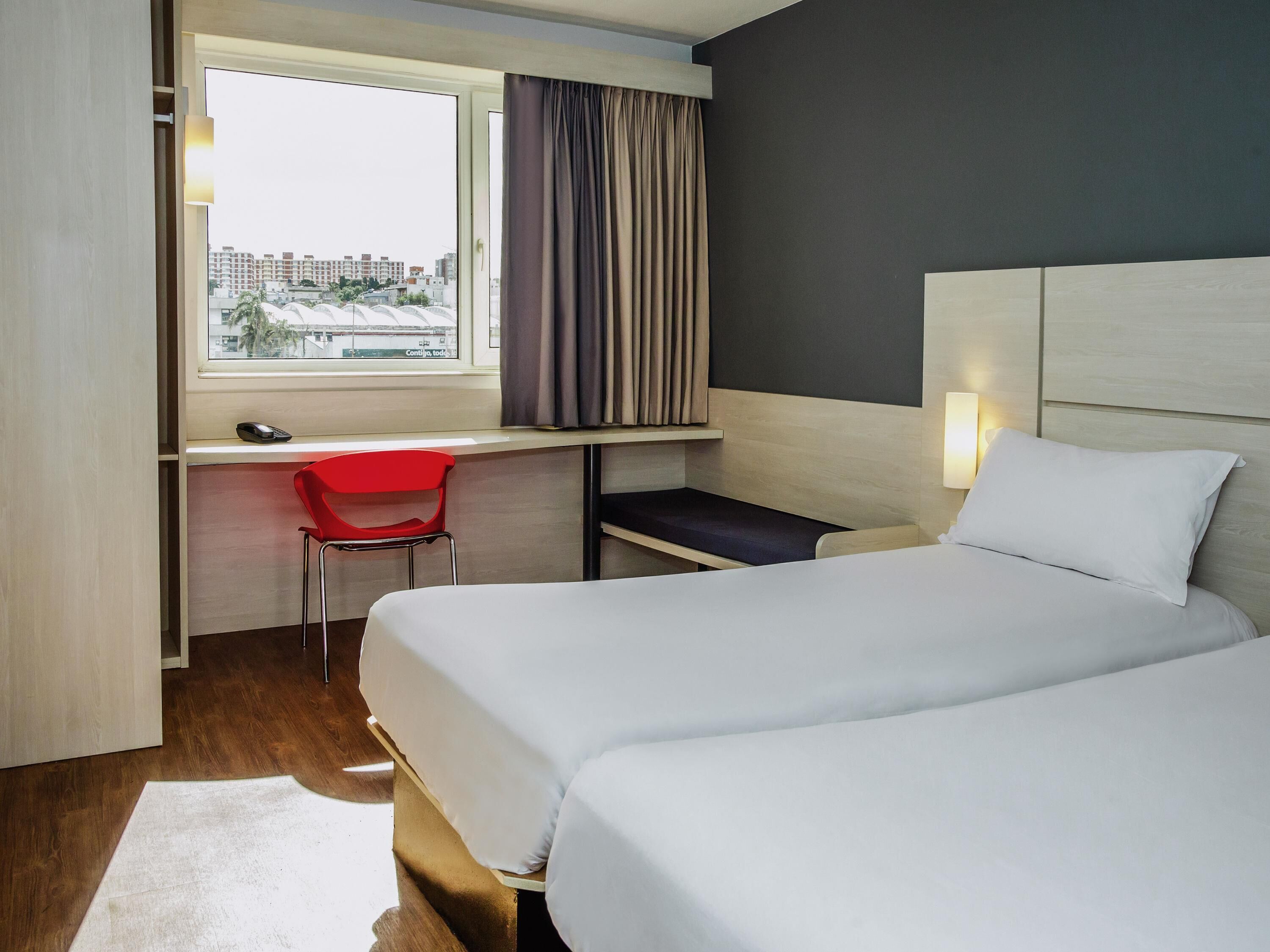 A hotel room with a bed, a desk with a chair, a window with curtains, and a city view.