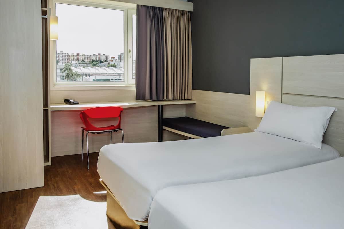 A hotel room with a bed, a desk with a chair, a window with curtains, and a city view.