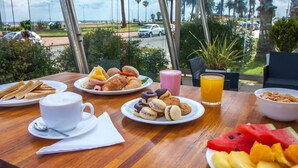 Una mesa de desayuno con platos de comida, una taza de café y un vaso de jugo, con vista a una calle con palmeras y coches estacionados.