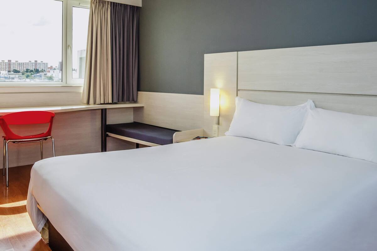 A hotel room with a bed, a desk, a chair, a window with curtains, and a nightstand with a lamp.