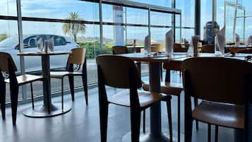 Um espaço de refeições interno com mesas, cadeiras, janelas amplas e vista para o exterior.