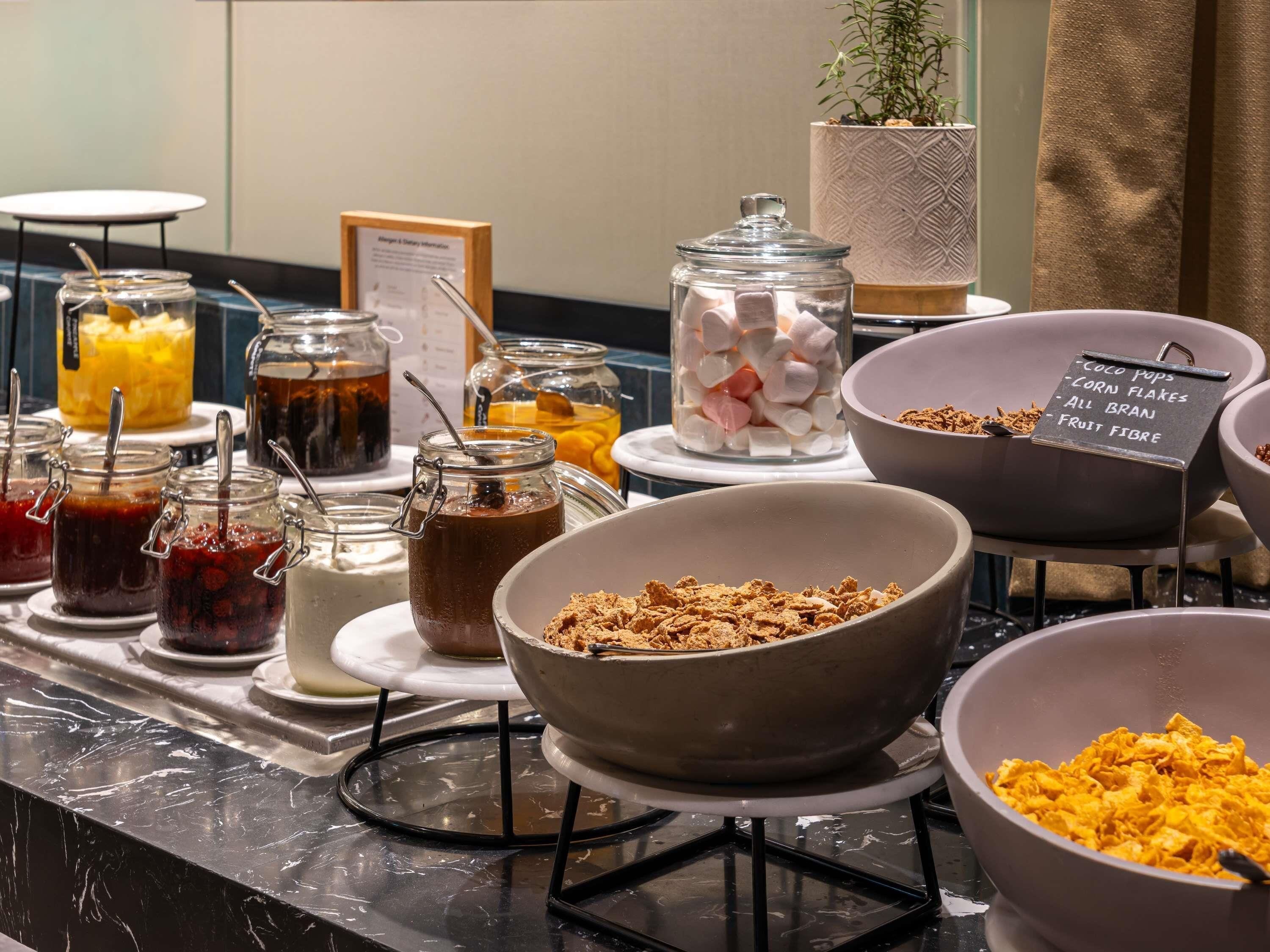 Un buffet con diversi cibi, tra cui vasetti di marmellata, una ciotola di cereali e un cartello che indica le opzioni disponibili.