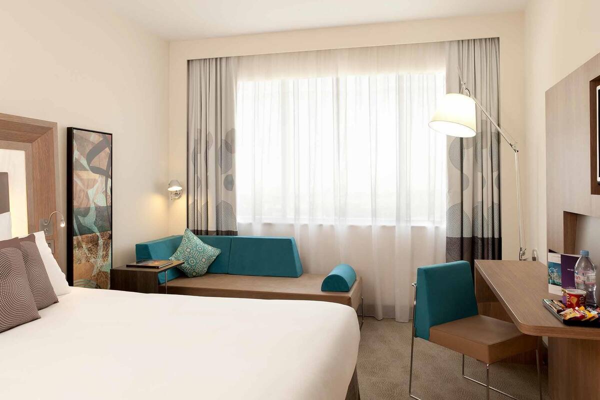 A hotel room with a bed, a desk, a chair, a sofa, and a large window with curtains.