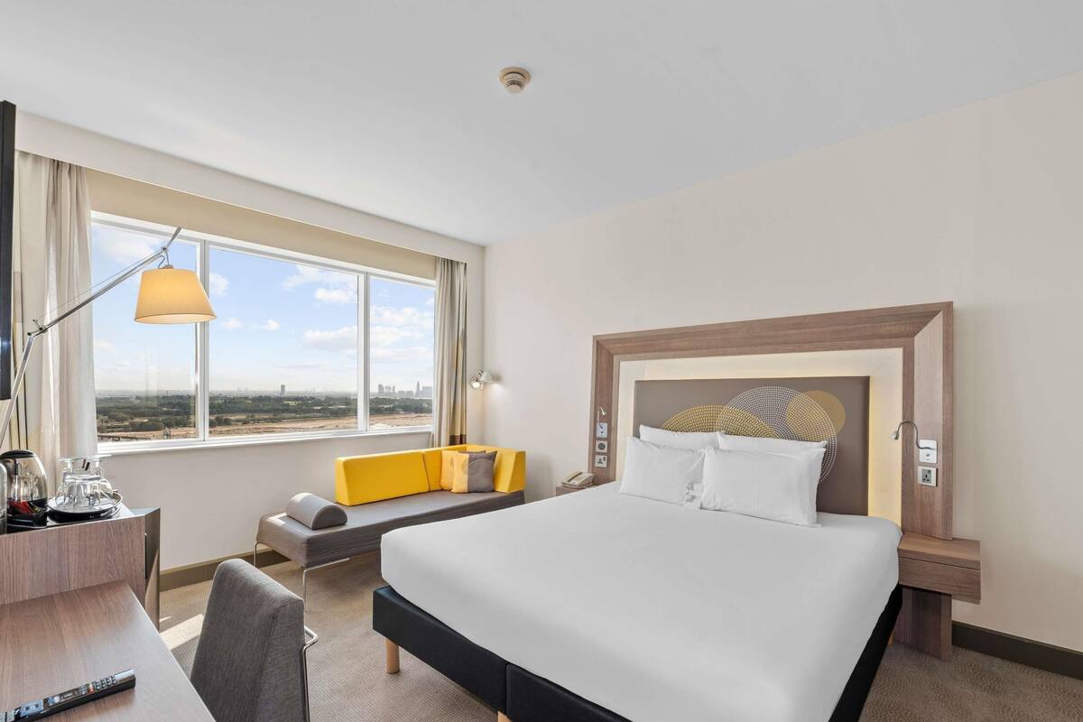 A hotel room with a large bed, a desk with a lamp, a sofa, and a view of the cityscape.