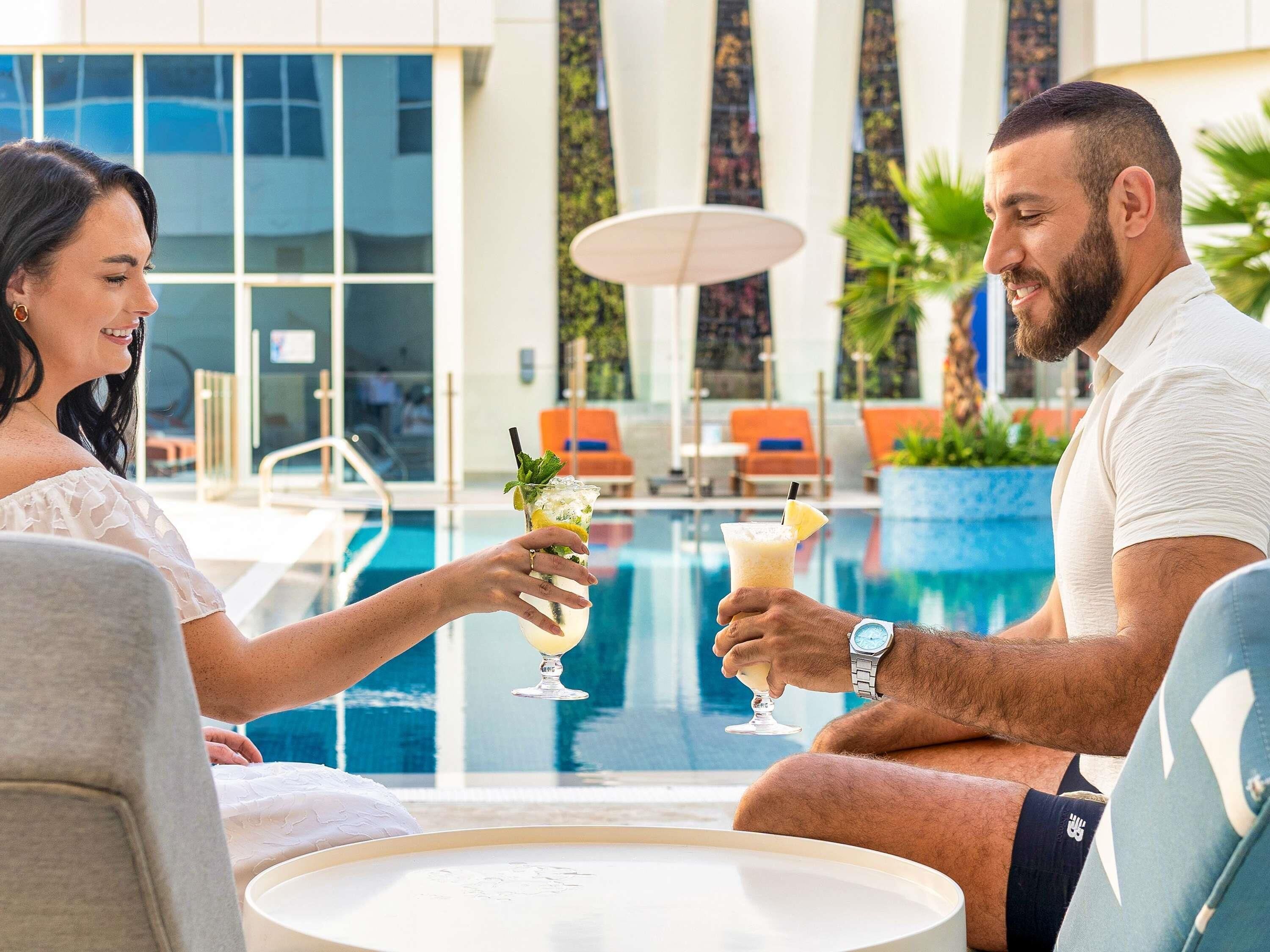 Un couple est assis au bord d’une piscine, des boissons à la main, entouré de chaises longues et d’un bâtiment moderne doté de grandes fenêtres.