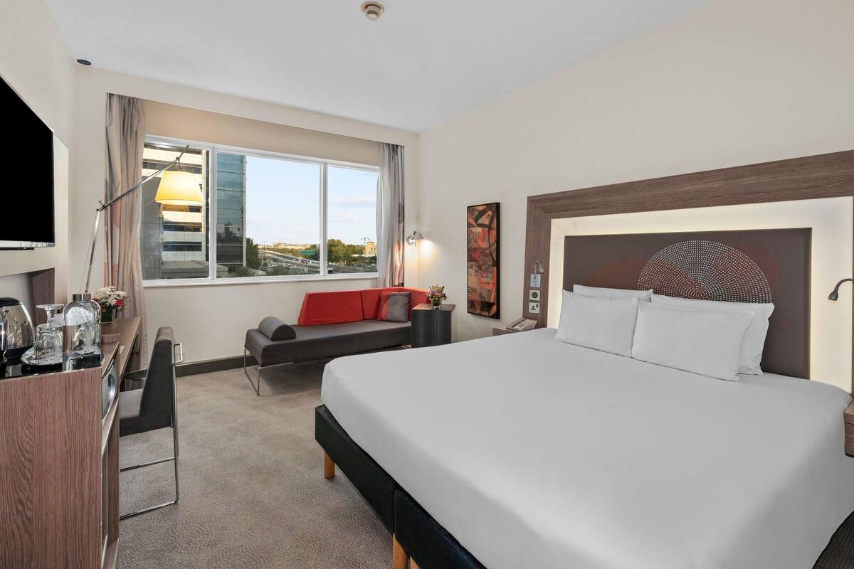 A hotel room with a large bed, a desk with a chair, a sofa, a TV, and a window with a city view.
