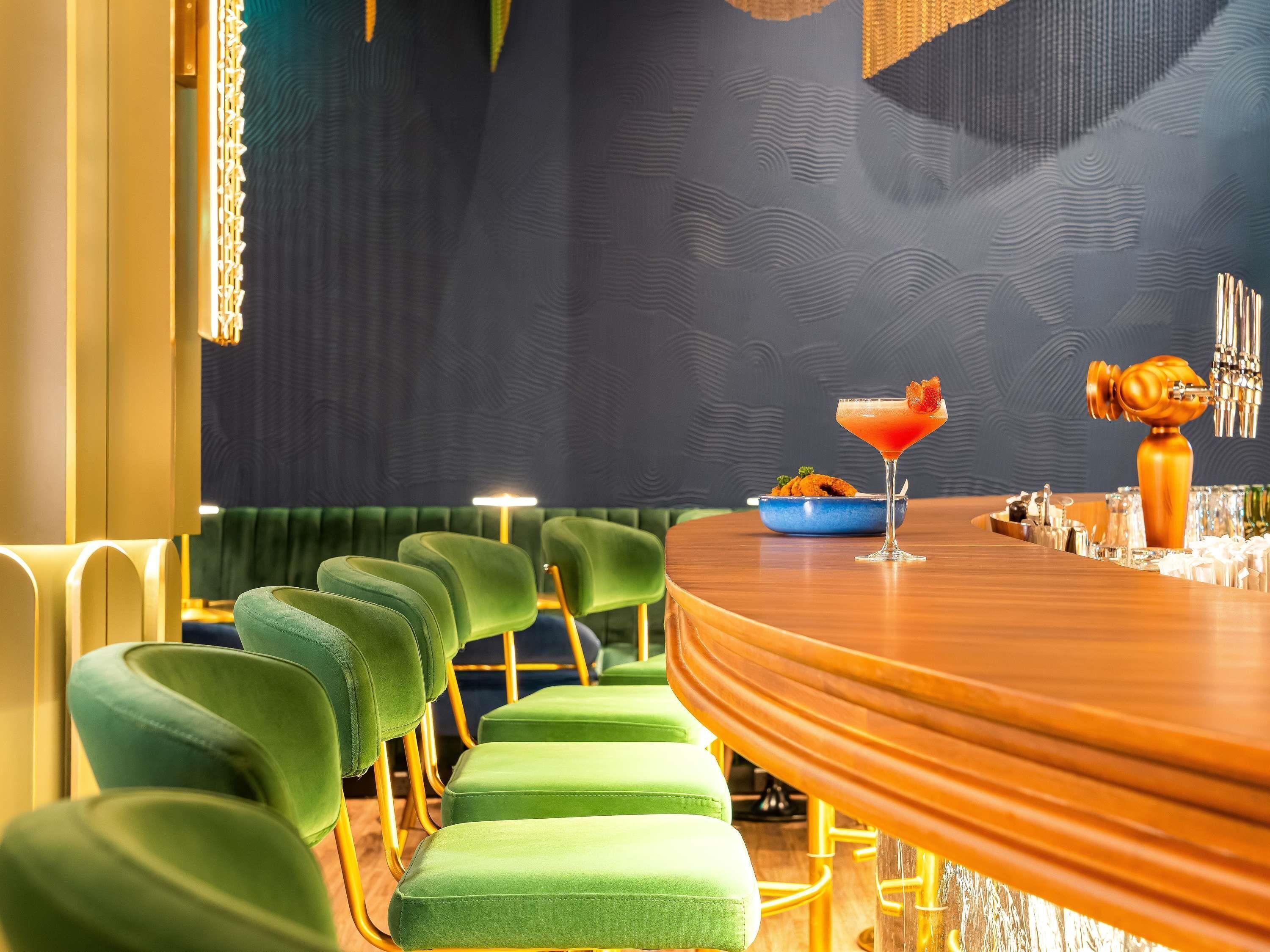 A bar area with a curved wooden counter, green upholstered bar stools, and a martini glass with a garnish.
