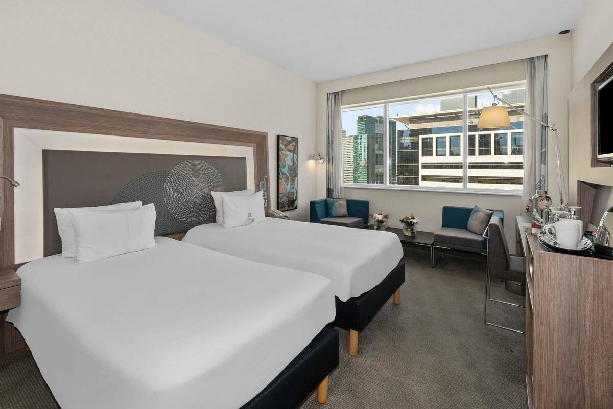 A hotel room with two beds, a desk, a chair, a TV, and a window with a city view.