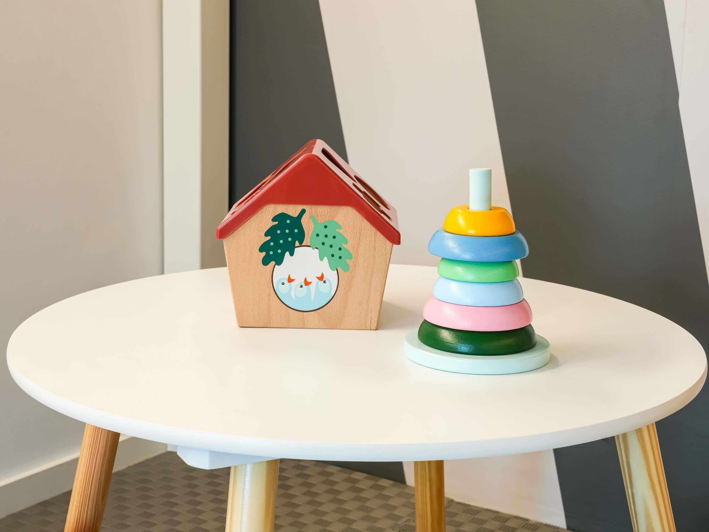 Una pequeña casa de madera con techo rojo y un juguete de anillos apilables de colores sobre una mesa blanca.