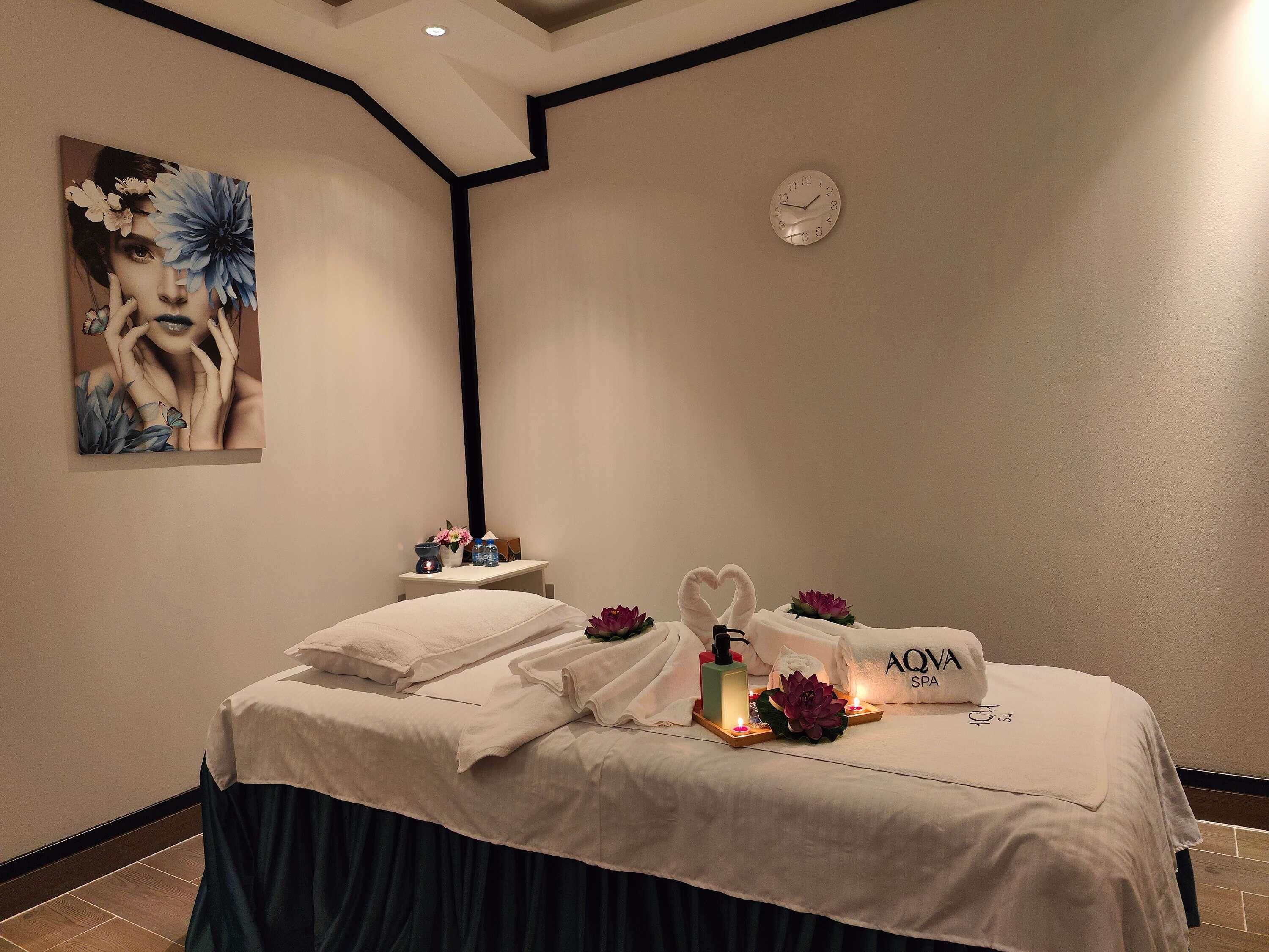 A spa room with a bed, towels, a clock, a painting, and a small table with a water bottle and a vase.