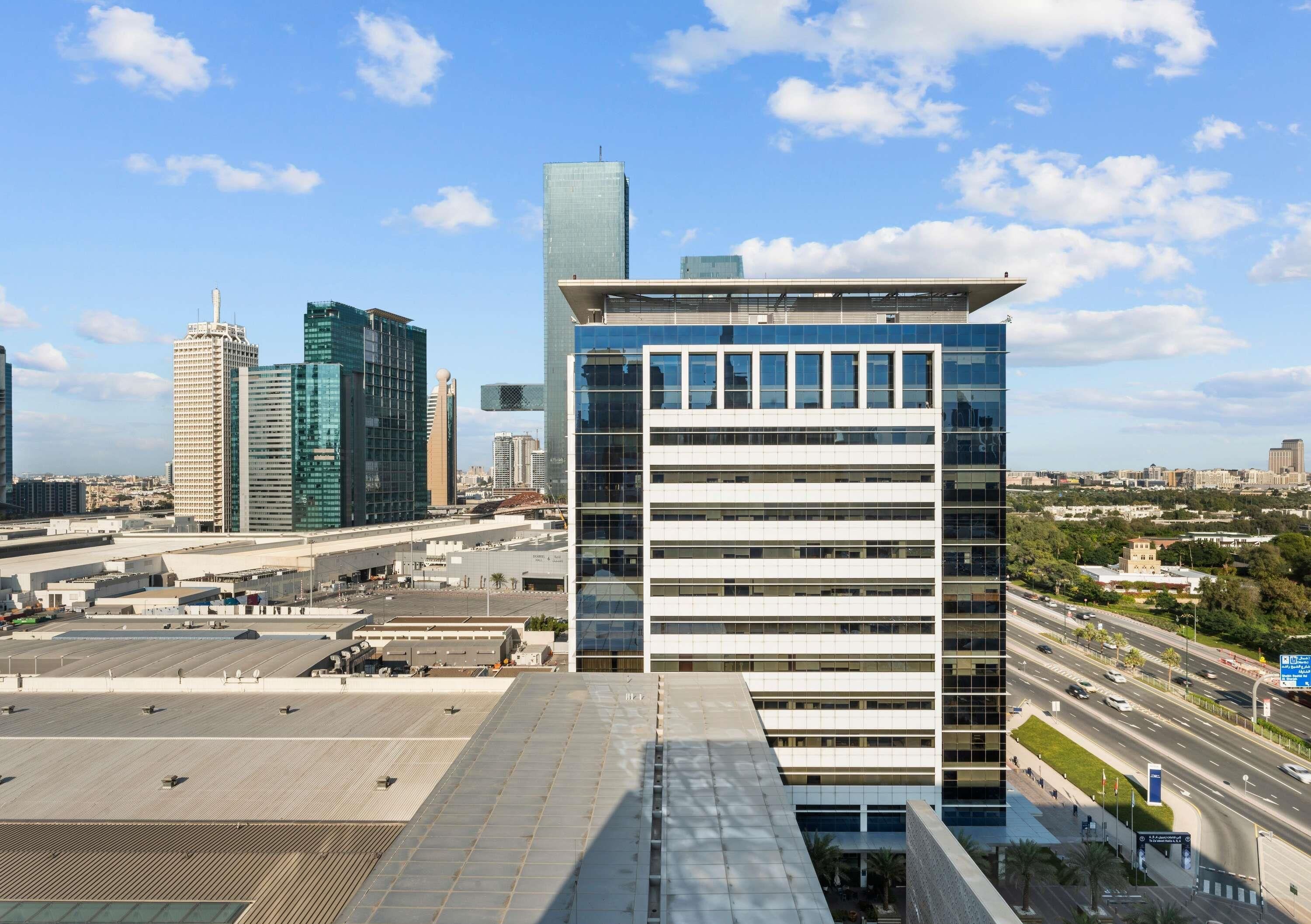 A modern building with a flat roof and multiple floors, surrounded by other high-rise buildings and a busy road.