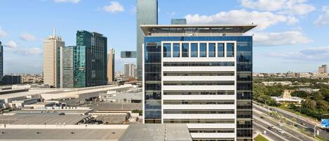 A modern building with a flat roof and multiple floors, surrounded by other high-rise buildings and a busy road.