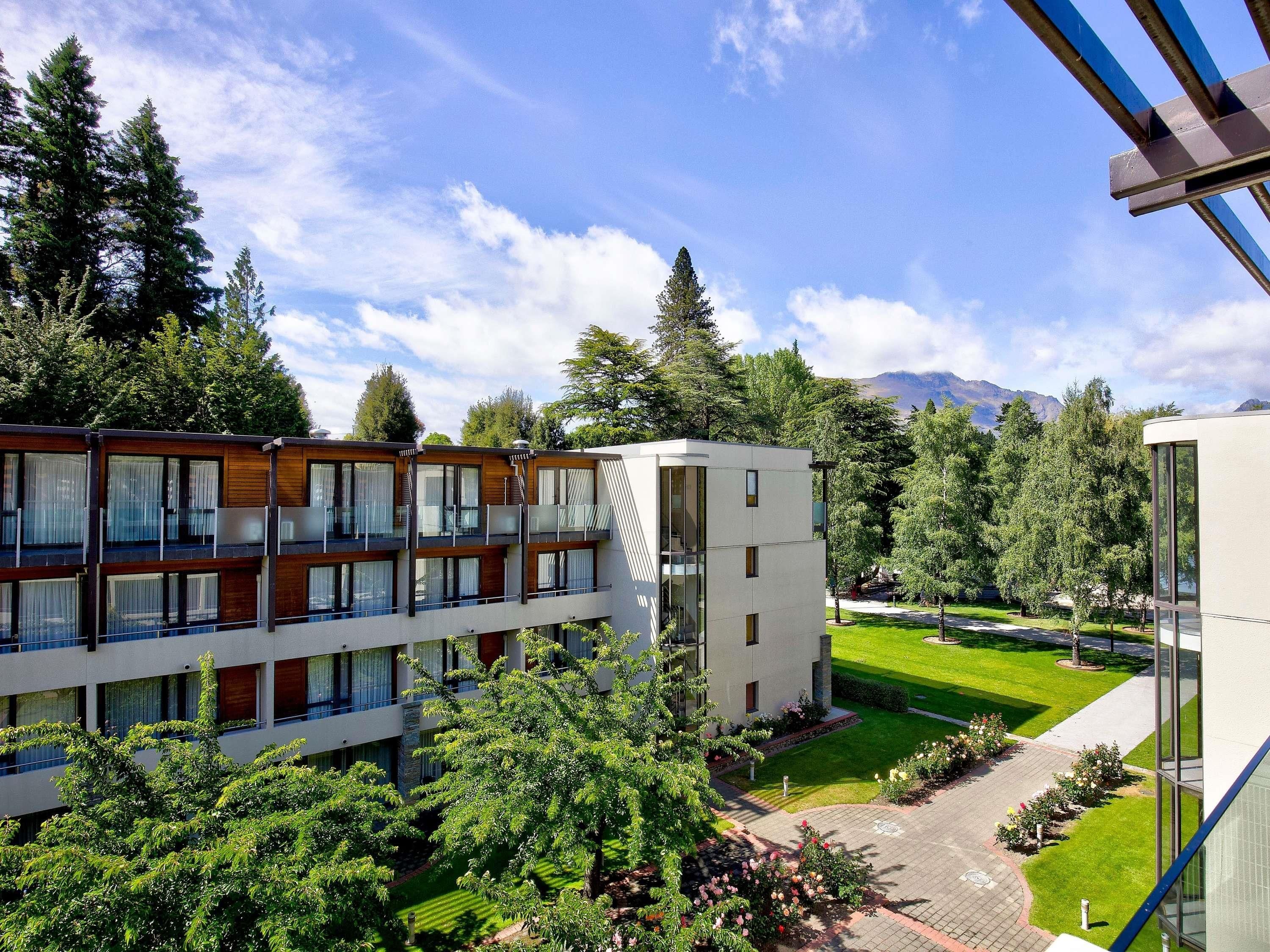 A modern multi-story building with a mix of wooden and white facades, surrounded by lush greenery and a well-maintained lawn.