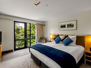 A hotel room with a large bed, a desk, a chair, a TV, a window with a view of trees, and a framed picture on the wall. - Novotel Queenstown Lakeside (Queenstown)