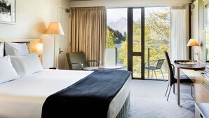 A hotel room with a large bed, two bedside lamps, a chair, a small table, and a view of mountains through a sliding glass door. - Novotel Queenstown Lakeside (Queenstown)