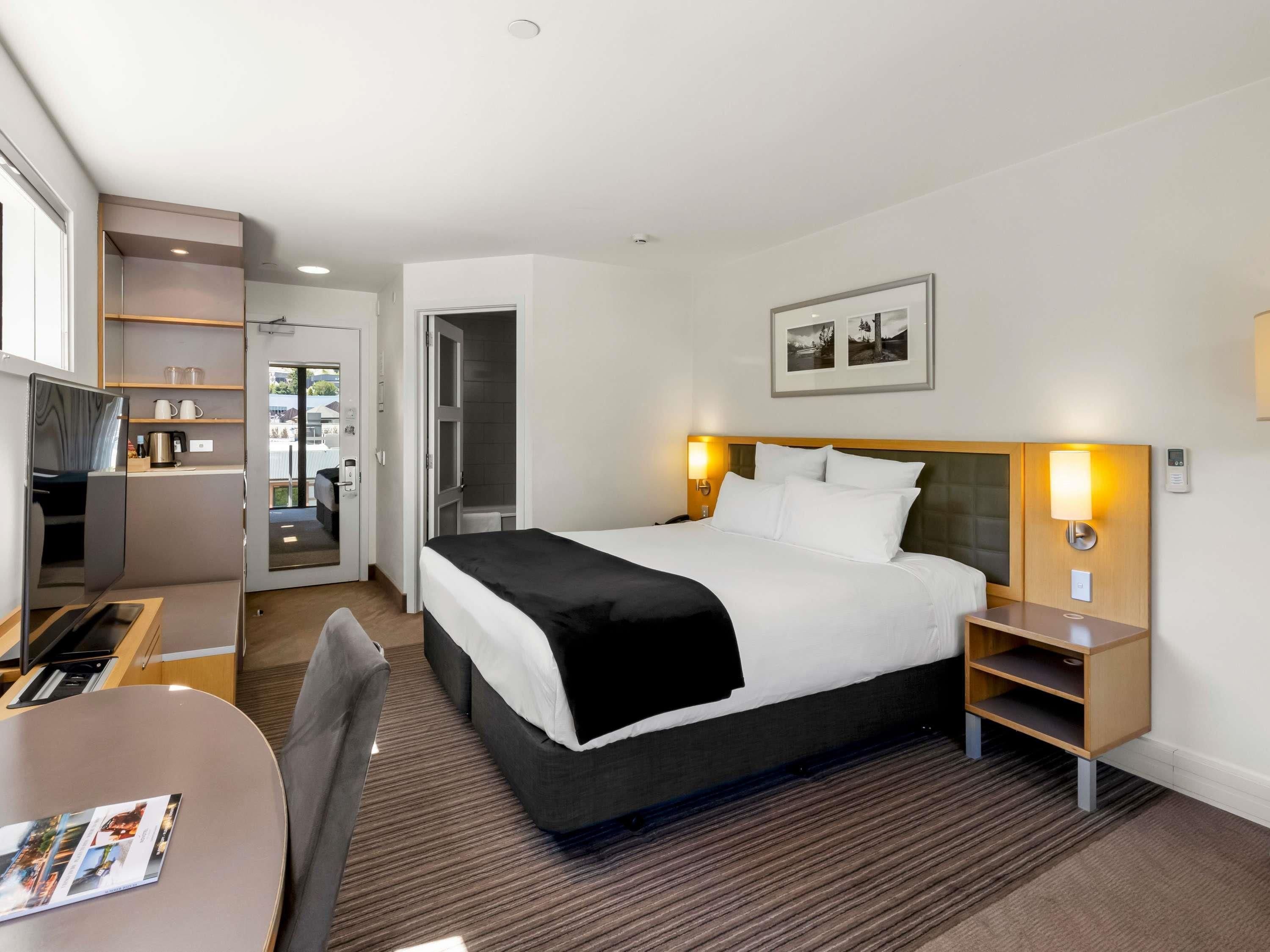 A hotel room with a bed, bedside tables, a desk, a chair, a TV, and a shower area.