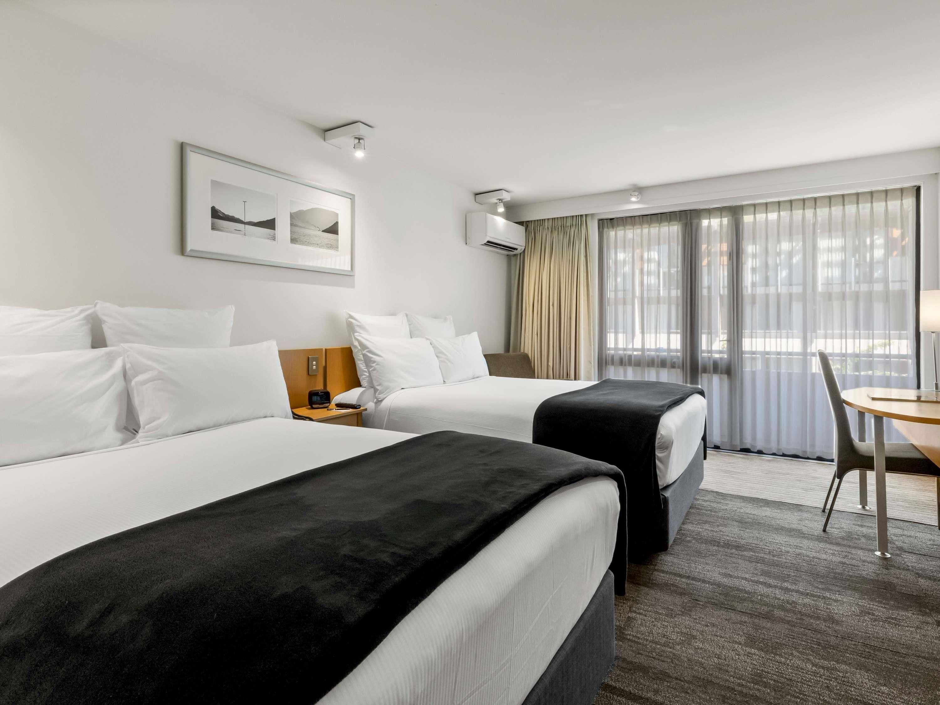 Habitación de hotel con dos camas, un escritorio y una silla. Cuenta con una ventana con cortinas, un cuadro en la pared y un equipo de aire acondicionado.