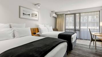 Habitación de hotel con dos camas, un escritorio y una silla. Cuenta con una ventana con cortinas, un cuadro en la pared y un equipo de aire acondicionado.