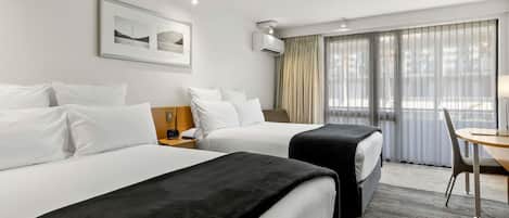 A hotel room with two beds, a desk, and a chair. There is a window with curtains, a framed picture on the wall, and an air conditioning unit.