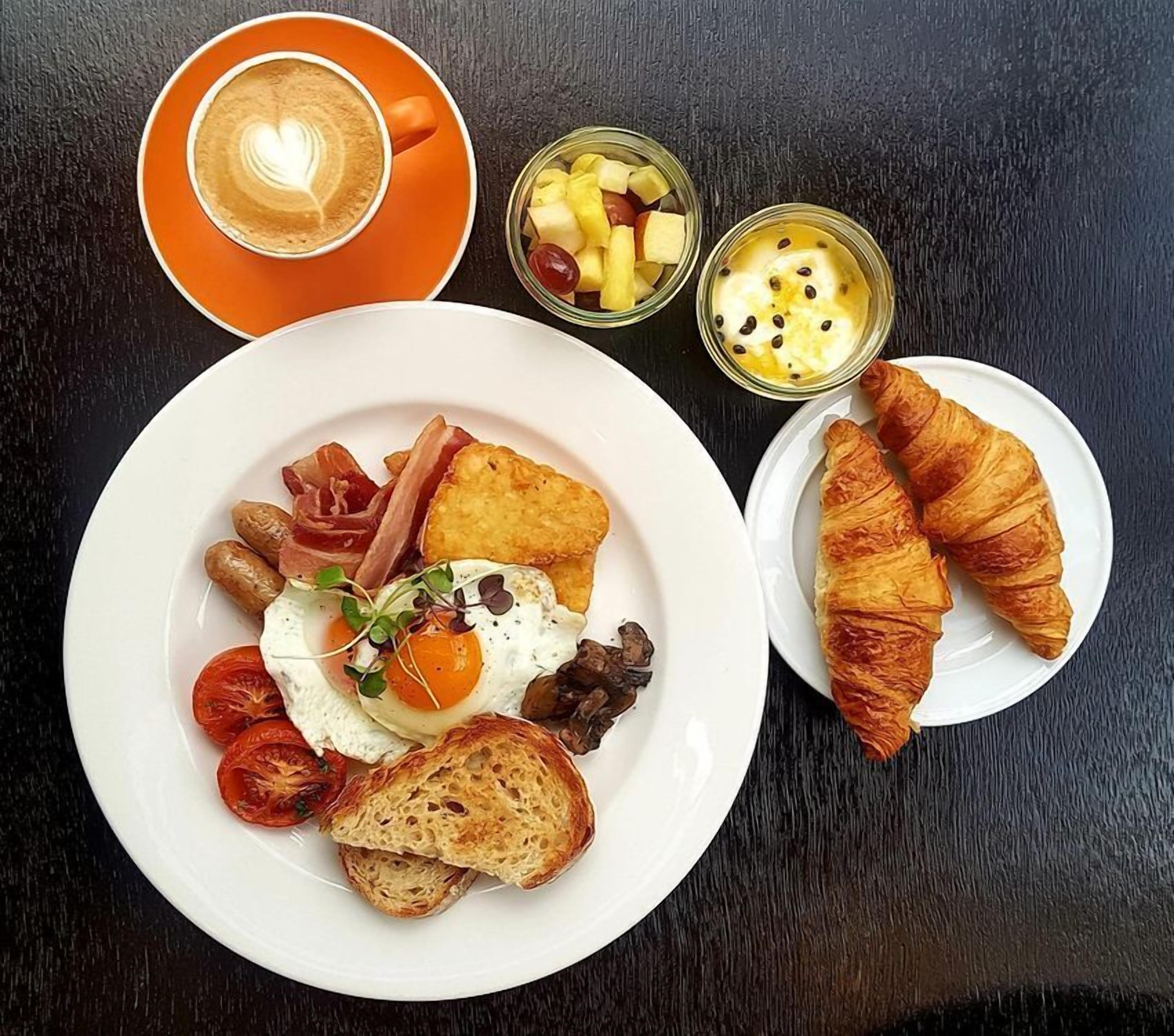 A breakfast plate with eggs, bacon, tomatoes, mushrooms, and toast, accompanied by a croissant and two small glasses of fruit and yogurt.