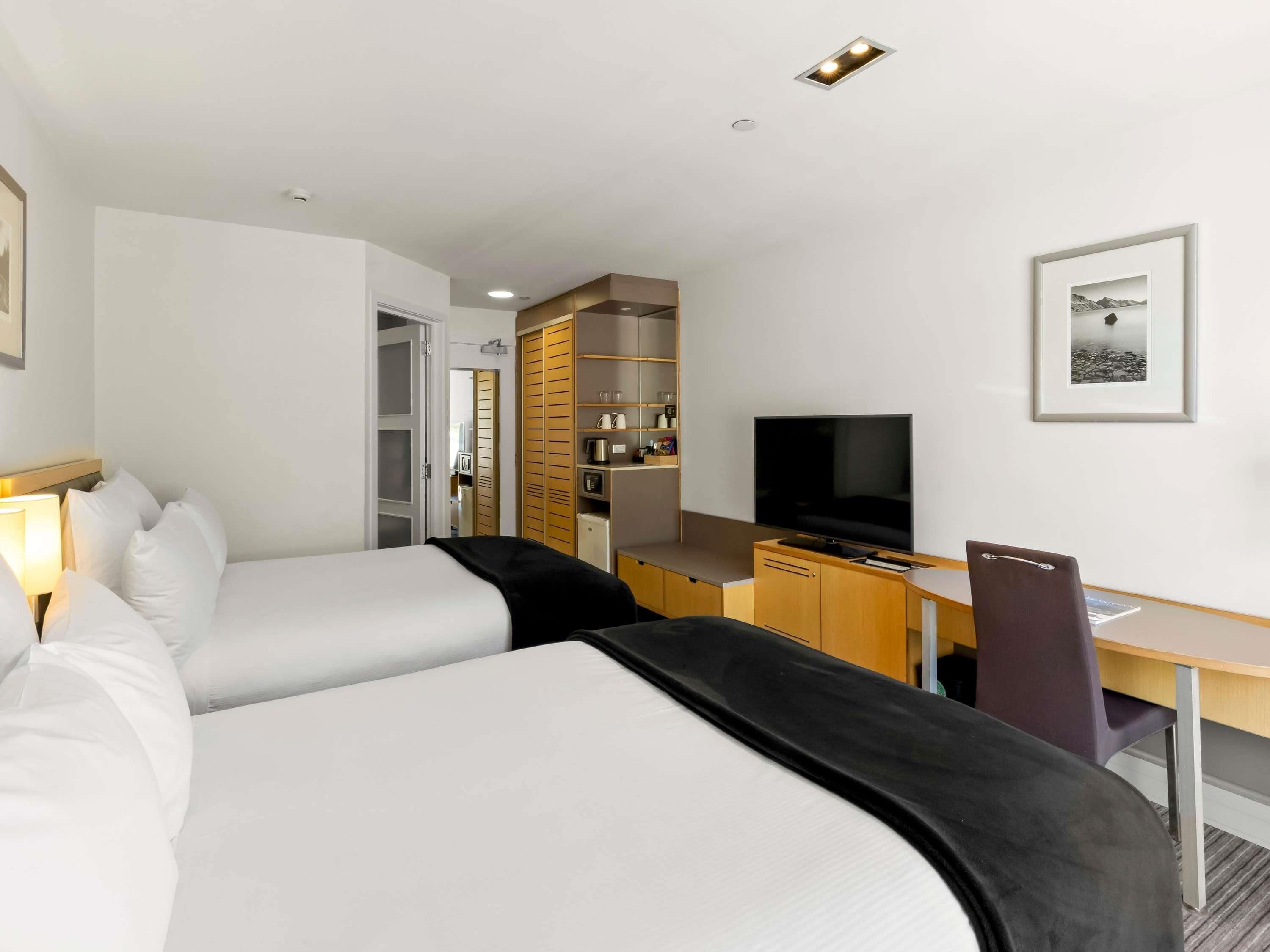 A hotel room with two beds, a desk with a TV, a chair, and a kitchenette.