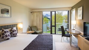 A hotel room with a bed, a desk, a chair, a TV, and a view of a lake and mountains. - Novotel Queenstown Lakeside (Queenstown)